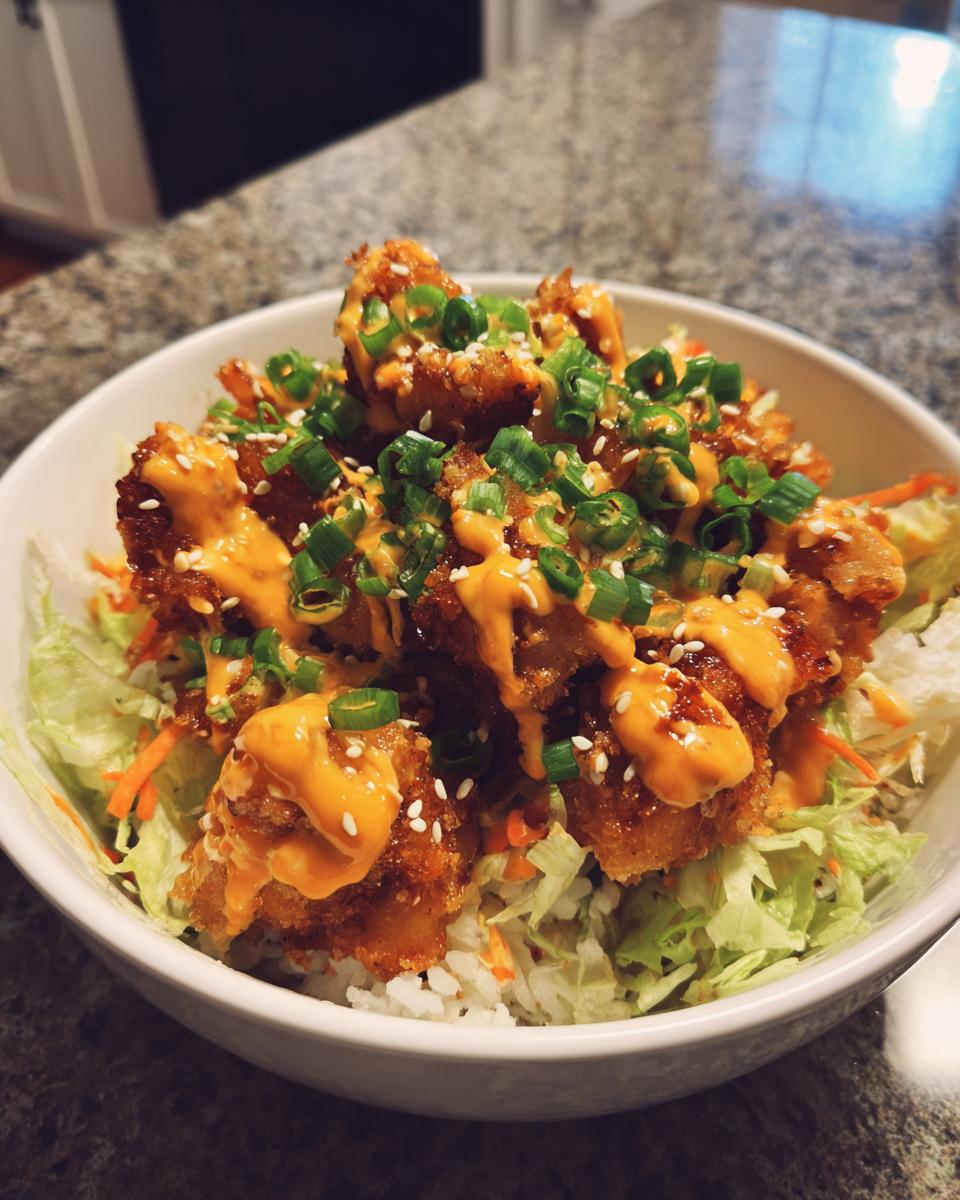 A close-up of a white bowl filled with rice, shredded lettuce, crispy Bang Bang Chicken pieces drizzled with orange sauce, and topped with green onions.