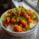 A close-up of a bowl filled with white rice, lettuce, and crispy bang bang chicken drizzled with orange sauce and topped with green onions.