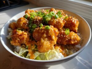 Close-up of crispy Bang Bang Chicken pieces drizzled with sauce, served over rice and shredded lettuce, topped with green onions and sesame seeds.