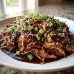 A plate of shredded crockpot teriyaki chicken coated in rich sauce, topped with green onions and sesame seeds.