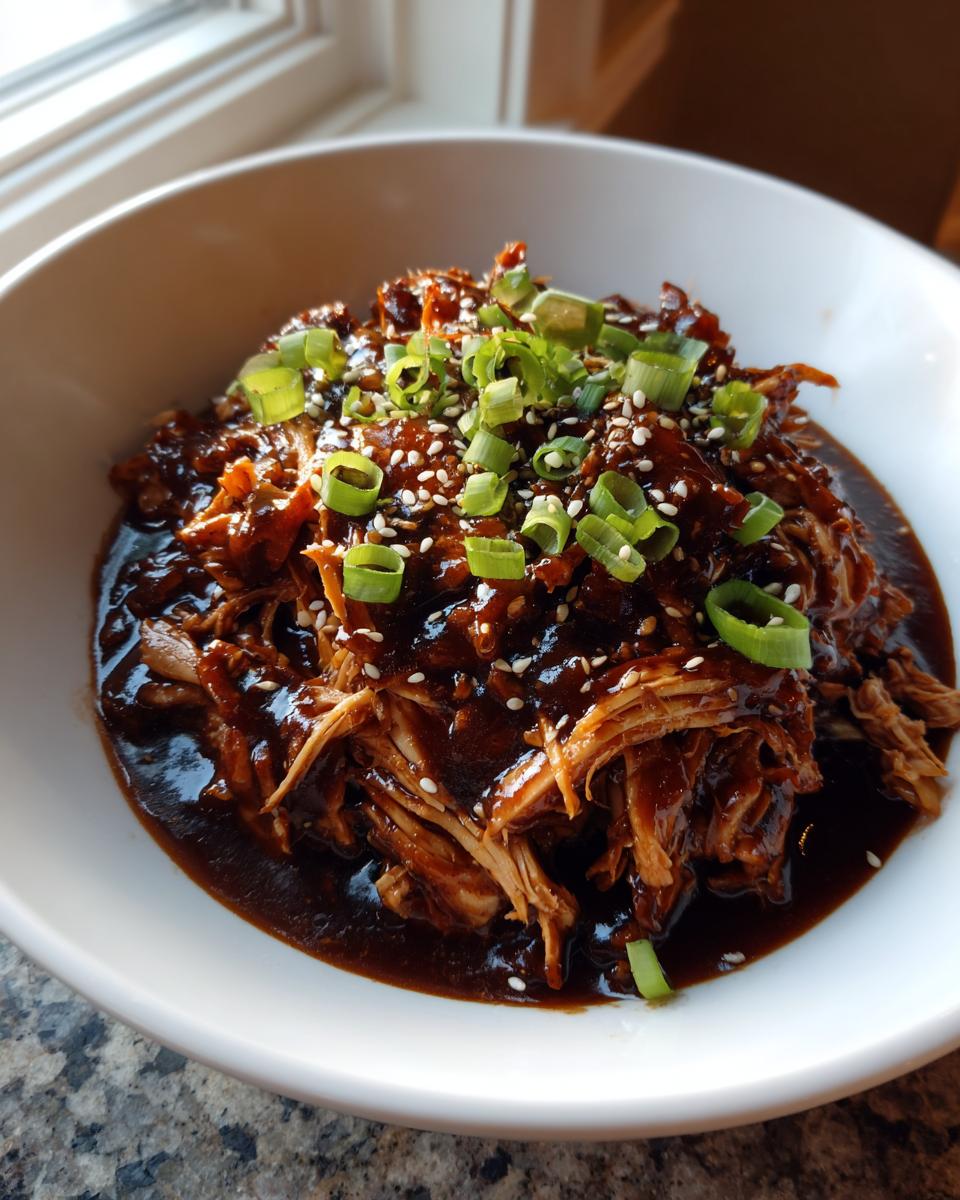 Close-up of shredded crockpot teriyaki chicken coated in dark sauce, topped with green onions and sesame seeds.