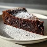 A close-up of a dense, dark slice of flourless chocolate cake dusted generously with powdered sugar on a light plate.