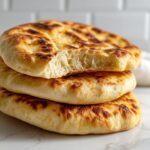 A stack of three golden brown, fluffy pita bread rounds, with the top one broken open to show the airy interior pocket.