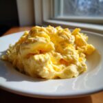 A close-up of perfectly cooked, fluffy scrambled eggs served on a white plate near a sunlit window.