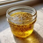 Close-up of homemade salad dressing in a small glass jar, showing oil and visible seeds floating on top.