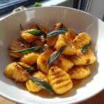 A white bowl filled with golden sweet potato gnocchi coated in a brown butter sauce and topped with crispy sage leaves.