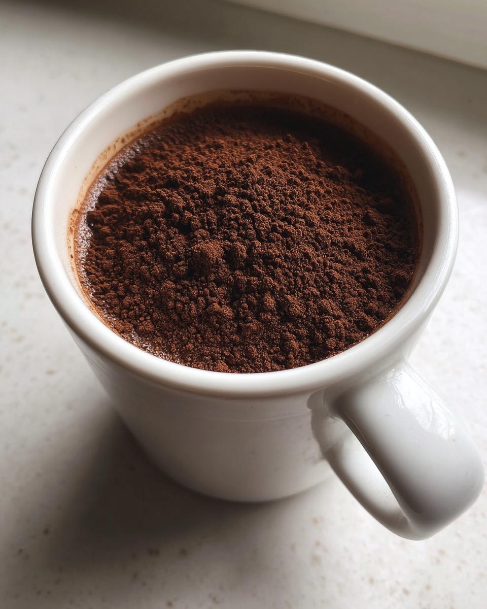 A close-up view of a white mug filled to the brim with rich, dark brown hot cocoa mix powder.