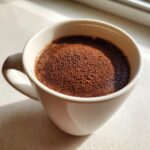 Close-up of a white mug filled with dark liquid, topped generously with rich cocoa powder from a hot cocoa mix.