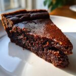A close-up of a moist, dark chocolate slice of fudge pie resting on a white plate.