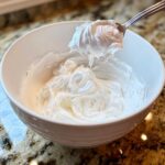 A spoonful of thick, bright white royal icing recipe mixture being lifted from a white bowl.