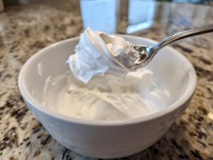 A spoonful of thick, white royal icing recipe mixture held above a white bowl showing perfect stiff peaks.