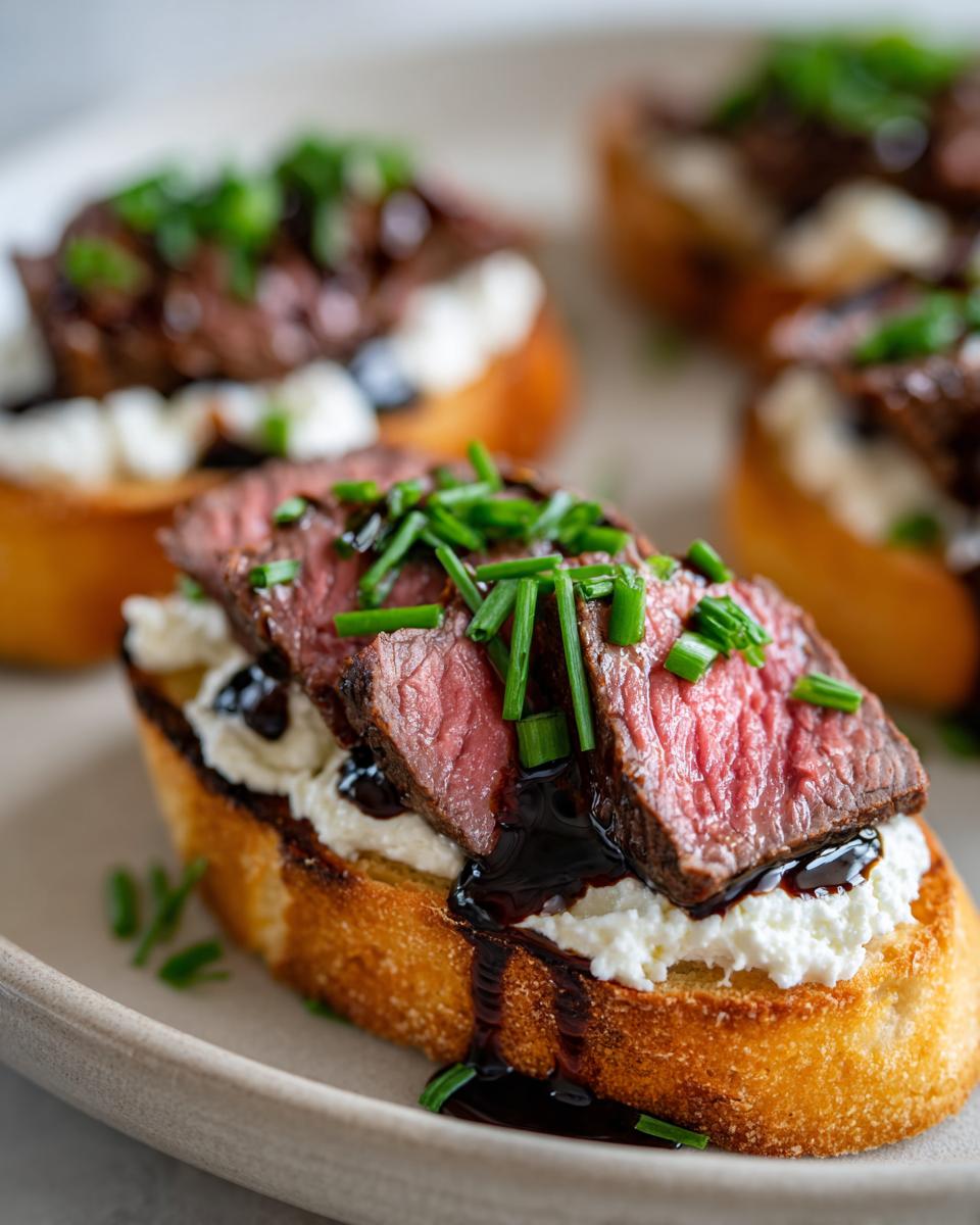 Close-up of a steak crostini bite topped with medium-rare steak, creamy cheese, balsamic glaze, and chives.