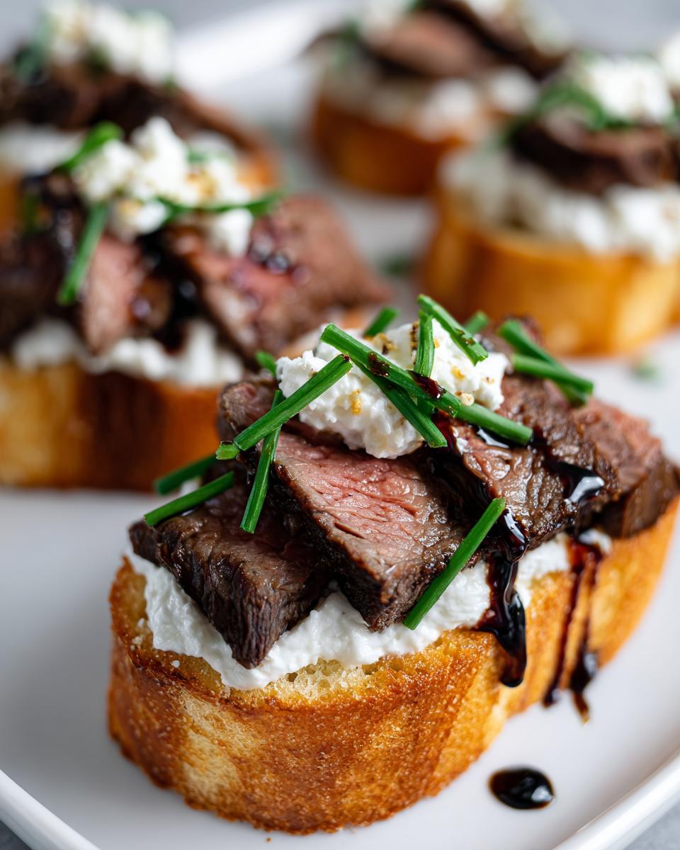 Close-up of a perfectly prepared steak crostini bite topped with medium-rare steak, creamy cheese, and fresh chives.