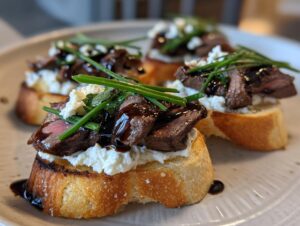 Four slices of steak crostini topped with creamy cheese, sliced steak, balsamic glaze, and fresh chives.