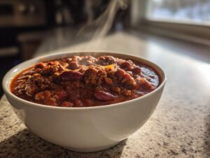 A steaming white bowl filled with rich, hearty high protein chili featuring ground meat and kidney beans, set on a countertop.