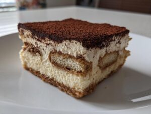 Close-up of a decadent slice of tiramisu cheesecake showing layers of cream, soaked ladyfingers, and a rich cocoa powder topping.