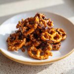 A close-up of a pile of crunchy toffee pretzels coated in a rich chocolate drizzle on a light plate.