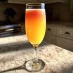 A vibrant, layered bellini recipe cocktail served in a champagne flute glass on a sunlit granite countertop.