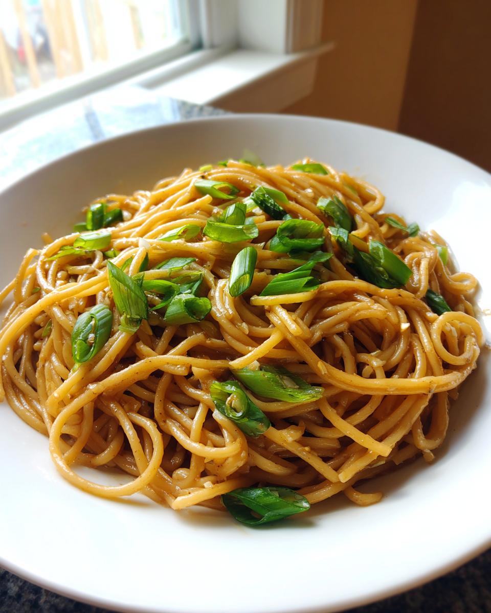 A close-up of a white bowl filled with glossy garlic noodles topped with fresh green onions.