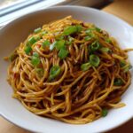 A close-up of a white bowl filled with glossy, saucy garlic noodles topped with fresh green onions.