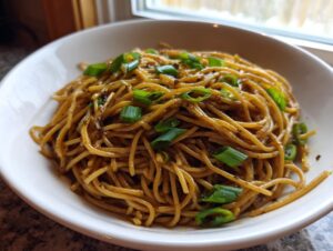 Close-up of savory brown garlic noodles tossed in sauce and topped with fresh green scallions in a white bowl.