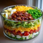 Close-up of a visually stunning 7 layer salad in a clear glass bowl, featuring layers of peas, cheese, bacon, and lettuce.