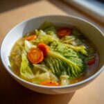 A close-up of vibrant cabbage soup featuring large pieces of green cabbage and bright orange carrot slices in a clear broth.
