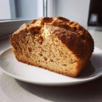 A rustic, golden-brown loaf of soda bread, sliced open to show the crumb texture, resting on a white plate.