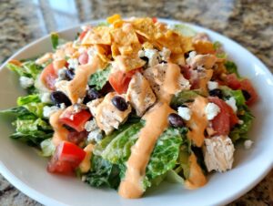 A close-up of a vibrant southwest salad featuring chopped chicken, black beans, tomatoes, cheese, and creamy dressing, topped with tortilla chips.
