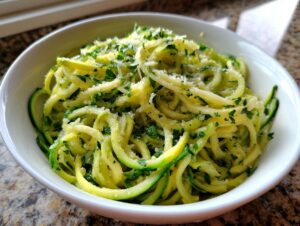 A close-up of a white bowl filled with fresh zucchini noodles tossed with herbs and grated cheese.
