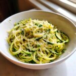Close-up of a white bowl filled with fresh zucchini noodles tossed in sauce and topped generously with grated Parmesan cheese and parsley.