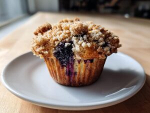 A moist blueberry muffin topped with a thick, sugary crumb topping, sitting on a small white plate.