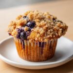 A close-up of a golden brown blueberry muffin featuring a generous, sugary crumb topping and visible blueberries.