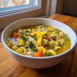 Close-up of a white bowl filled with rich chicken noodle soup, featuring egg noodles, shredded chicken, carrots, and parsley.