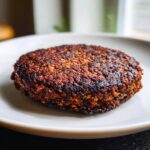 Close-up of a perfectly seared, dark brown black bean burger patty resting on a white plate.
