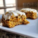A close-up of a soft carrot cake cookie, broken in half, showing shredded carrots and topped with thick cream cheese frosting.