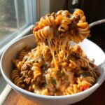 A fork lifts a cheesy portion of taco pasta out of a white bowl, showing long cheese pulls.