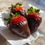 Three fresh, glossy chocolate covered strawberries sitting on a white napkin in bright sunlight.