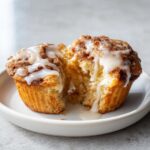 A cinnamon roll muffin split in half showing the soft interior and drizzled with white icing.