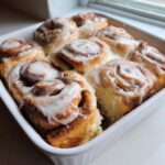 Close-up of gooey, freshly baked cinnamon rolls recipe topped with thick white icing in a white baking dish.