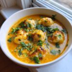 A close-up of a white bowl filled with rich, orange coconut curry soup with dumplings, garnished heavily with fresh cilantro.