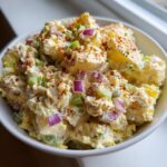 Close-up of creamy potato salad mixed with red onion and celery, topped with paprika.
