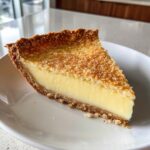 A thick slice of creamy buttermilk pie with a golden, slightly crumbly top crust, served on a white plate.