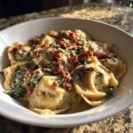 Close-up of creamy tuscan ravioli featuring spinach and sun-dried tomatoes in a white bowl.