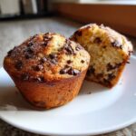 Two fluffy chocolate chip muffins on a white plate, one cut in half showing the moist interior.