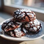A stack of four rich, dark chocolate crinkle cookies heavily dusted with white powdered sugar.