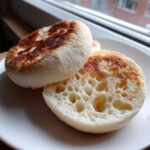 Two homemade english muffins, one split open showing the classic open crumb structure, lightly toasted on a white plate.