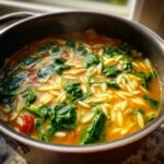 Close-up of steaming italian orzo spinach soup filled with orzo pasta, bright green spinach, and tomatoes in a dark pot.