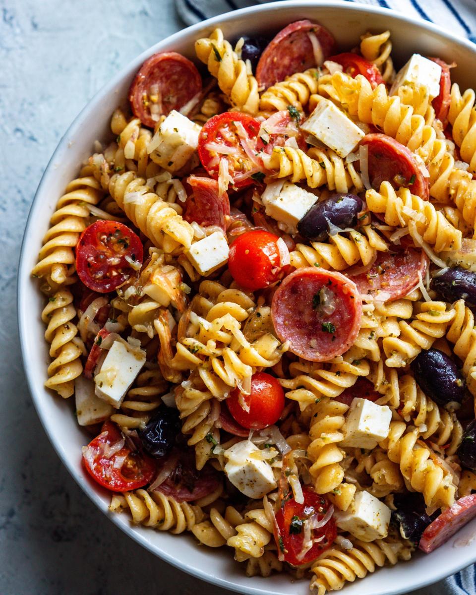 Close-up overhead view of a bowl filled with Italian pasta salad featuring rotini, salami, mozzarella cubes, cherry tomatoes, and olives.