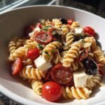 A close-up view of a bowl filled with vibrant italian pasta salad featuring fusilli, pepperoni, cherry tomatoes, olives, and mozzarella cubes.